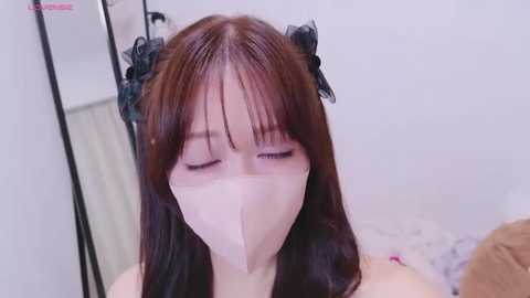 Media: A video of a young East Asian woman with long, straight brown hair, wearing black lace headbands, and a pink face mask, sitting indoors with white walls and a mirror visible.