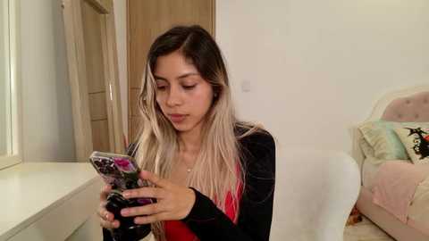 Media: Video of a young woman with long, wavy blonde hair, light skin, and a slender physique, wearing a black velvet jacket over a red top, sitting on a white chair, looking at a phone with a floral case, in a minimalist, brightly lit bedroom.