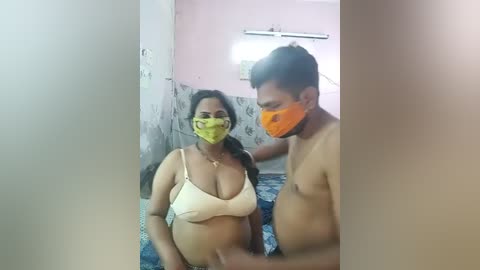 Media: A video captures a pregnant woman in a yellow mask, wearing a beige bra, and a man in an orange mask, both standing in a small, dimly-lit room with a blue-patterned bed.