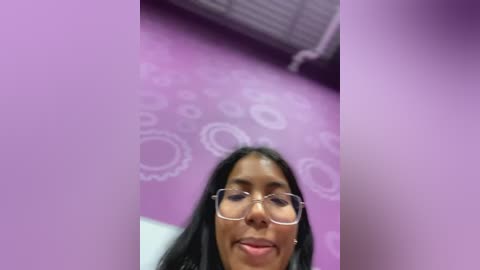 Media: Video of a woman with long black hair and glasses, standing in a purple room with white circular designs on the wall. The image is slightly blurry and has a purple filter effect.