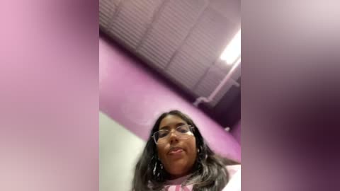 Media: A video of a smiling South Asian woman with long black hair, wearing glasses and a pink shirt, taken from a low angle, in a purple-painted room with a grid ceiling.