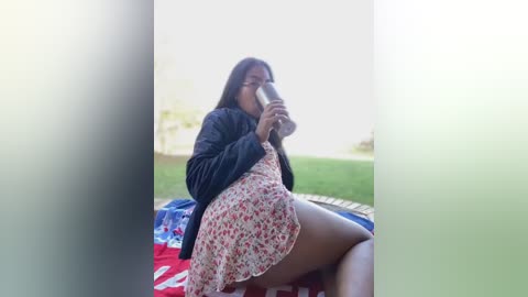 Media: A young Black woman with long hair, wearing a floral dress and black jacket, drinks from a water bottle while sitting on a red and white picnic blanket in a grassy park.