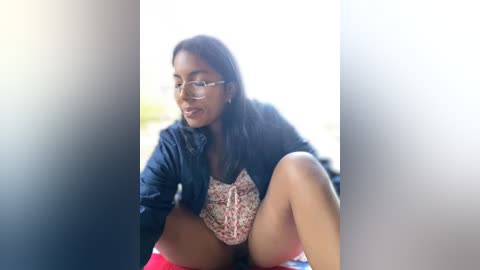 Media: Video of a smiling, dark-skinned woman with glasses, wearing a floral top and shorts, sitting outdoors on a blanket with blurred background.