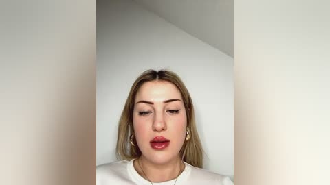 Media: Video of a young woman with fair skin and long blonde hair, wearing white clothing, standing against a plain white wall. She has a neutral expression and full lips.