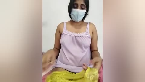 Media: A video of a South Asian woman with medium skin tone, wearing a light pink tank top, yellow shorts, and a blue surgical mask, sitting indoors with a plain background.