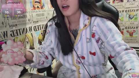 Media: Video of a young woman in a striped pajama top, holding a pink fluffy toy, in a cluttered room with newspapers and cartoon posters.