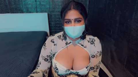 Media: A video of a woman with medium skin tone, wearing a floral-patterned white blouse, revealing cleavage, and a blue surgical mask. She has dark hair tied back and sits on a bed with dark bedding and a brick wall background.