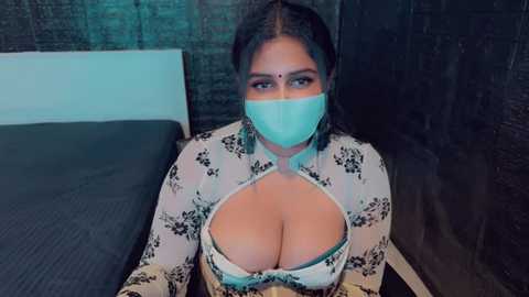 Media: Video of a woman with medium skin tone, wearing a white floral blouse with a large keyhole exposing her ample cleavage and a teal face mask. She has long dark hair and sits on a bed with a dark grey blanket.