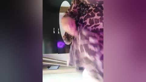 Media: Video of a person wearing a leopard print coat, standing in a dimly lit room with purple lighting. Background shows a wooden staircase and a smartphone on the floor.