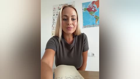 Media: Video of a blonde woman with straight hair, wearing a gray t-shirt, sitting at a desk, holding an open book, in a classroom with colorful world maps and educational charts on the wall.