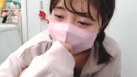 Media: Video of an Asian woman with medium-length black hair, wearing a light pink surgical mask, looking down, in a white room with stuffed toys.