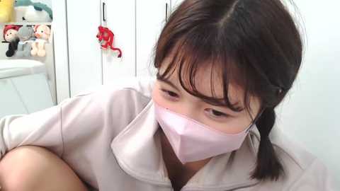 Media: A video of an Asian woman with dark hair in braids wearing a pink surgical mask and a light beige medical gown, standing in a white room filled with stuffed animals and medical supplies.