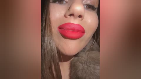 Media: A close-up video of a woman with light brown skin and long, straight, dark brown hair. Her full, glossy red lips are prominently featured, and she wears large silver hoop earrings. The background is blurred with a warm, reddish hue.