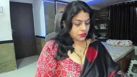 Media: Video of a South Asian woman with long black hair, fair skin, wearing a red patterned saree, sitting in a modern bedroom with a bed and wooden furniture.