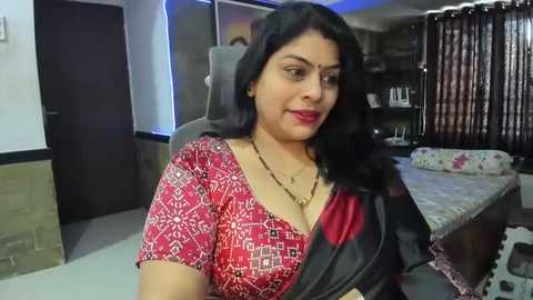 Media: Video of a smiling South Asian woman with long black hair, wearing a red-patterned blouse and black sari, in a modern, dimly lit bedroom with a bed, bookshelf, and curtains.