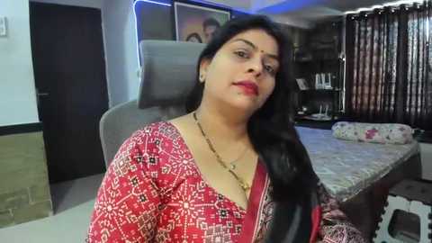 Media: Video of a South Asian woman with long black hair and fair skin, wearing a red patterned sari, seated in a modern bedroom with a TV, bookshelf, and bed.