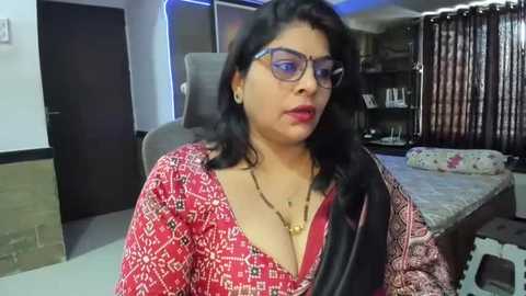 Media: Video of a middle-aged South Asian woman with dark hair, wearing glasses and a red patterned blouse with black trim, sitting in a dimly lit living room with a bed, shelves, and curtains in the background.