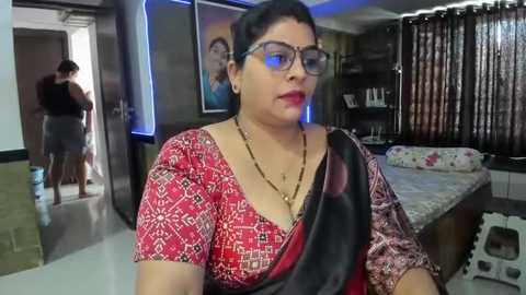 Media: Video of a middle-aged woman with short dark hair, wearing glasses and a red and black patterned sari, standing in a modern bedroom with a large bed, curtains, and a man in the background.