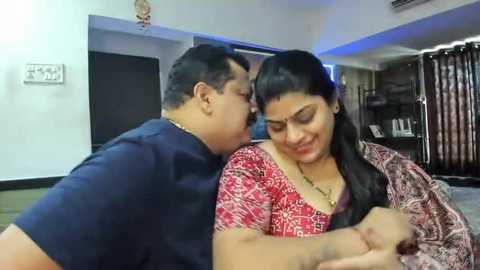 Media: Video of an Indian couple, a man in a blue shirt and woman in a red sari, hugging in a modern living room with a flat-screen TV and dark curtains.