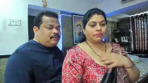 Media: A video of an elderly man with a mustache in a blue shirt and a young woman with dark hair and a red-patterned blouse, standing indoors near framed photos on a wall.