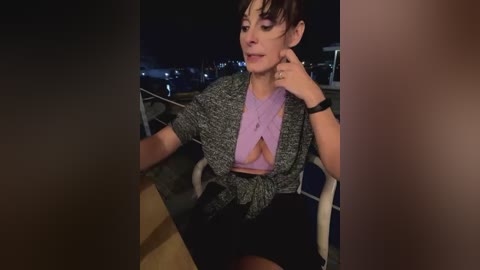 Media: Video of a woman with short dark hair, wearing a purple top and gray cardigan, sitting on a balcony at night.