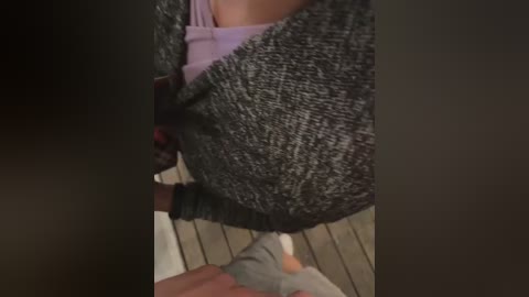 Media: Video of a woman's torso and lower legs, wearing a gray knit sweater over a lavender shirt and gray socks. She stands on a wooden deck, with a dark background.