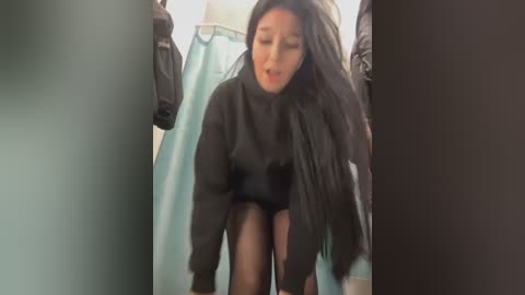 Media: A video of a young East Asian woman with long black hair, wearing a black sweater and sheer black tights, bending over in a public restroom. The background includes a blue curtain and other clothing items.