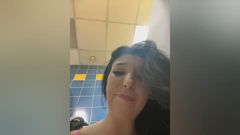 Media: Video of a woman with dark hair and light skin in a bathroom with blue and yellow tiled walls, smiling slightly.