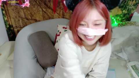 Media: Video of a smiling Asian woman with red hair, wearing a pink face mask, and a white long-sleeve shirt, sitting on a grey couch. Background includes a textured wall and colorful decor.