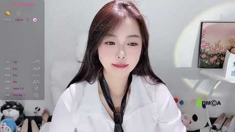 Media: A video of a young Asian woman with straight black hair and fair skin, wearing a white lab coat and black tie, standing in a bright, minimalist room with a large framed photo of flowers on the right wall.