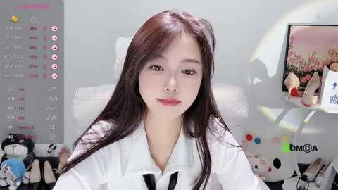 Media: Video of an East Asian woman with long black hair, light skin, and red lips, wearing a white shirt, seated in a bright room with a white bed, stuffed animals, and a screen displaying a nature scene.