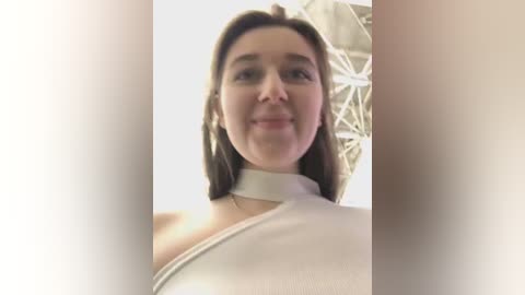 Media: Video of a smiling, light-skinned woman with shoulder-length brown hair, wearing a white, high-necked dress, standing indoors near a white, geometric-patterned wall.