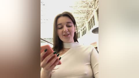 Media: Video of a young woman with light skin, straight brown hair, and green nail polish, wearing a white top, smiling at a smartphone in a modern, glass-walled indoor setting with industrial architecture.