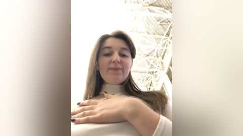 Media: A video of a young woman with light skin and straight brown hair, wearing a beige sweater, standing in a bright, modern indoor setting with a white, geometrically designed ceiling.