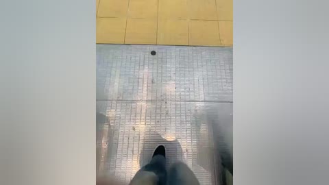 Media: Video of a person's feet on a glass floor, looking down. The floor is transparent, revealing a tiled wooden floor beneath. The scene is brightly lit, highlighting the textures and reflections.