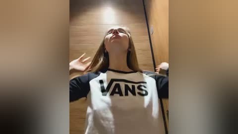 Media: A video of a young woman with long blonde hair, wearing a black and white Vans T-shirt, standing in a hallway with a wooden floor, looking up with a joyful expression.