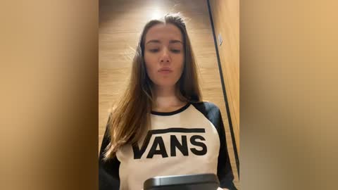 Media: Video of a young woman with long brown hair, wearing a white and black Vans t-shirt, standing in a wooden hallway with beige walls.