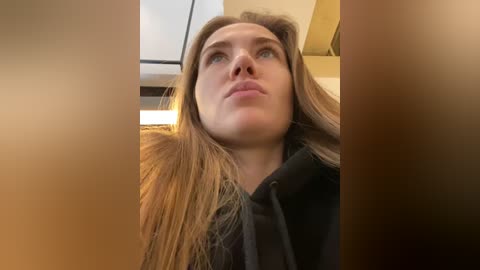 Media: Video of a woman with long, straight brown hair and fair skin, wearing a black jacket, looking upward with a contemplative expression. Background shows a blurred, beige and white interior setting.