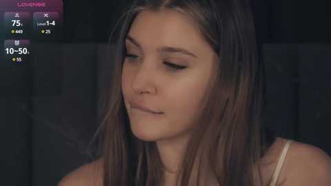 Media: Video of a young woman with light brown hair, wearing a white tank top, in a dimly lit room with a dark background.