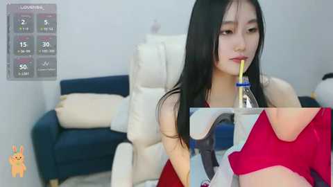 Media: Video of an East Asian woman with long black hair, wearing a red top, drinking from a plastic bottle, sitting on a white couch with blue cushions in a modern living room.