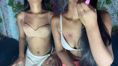 Media: Video of two slender women with medium skin tones, wearing lingerie. One has a beige bra, the other a white bra, both with red marks on their nipples. Background features floral patterns and a green plant.