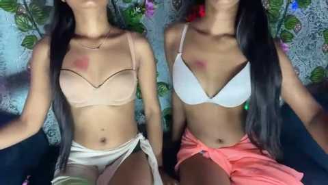 Media: Video of two young, slender, tan-skinned women with long black hair, wearing matching beige and white bras, and orange sarongs, seated against a floral-patterned background.