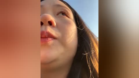 Media: Video of a young Asian woman with smooth, light brown skin, long dark hair, and a closed-lip smile, captured from a low angle, with a warm, golden glow and a blurred background.