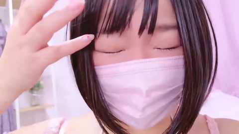 Media: A close-up video of an Asian woman with straight black hair and bangs, wearing a white face mask, having her hair combed by another hand.