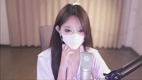 Media: Video of an Asian woman with long brown hair, wearing a white mask and shirt, sitting in front of a microphone in a dimly lit room with beige curtains.