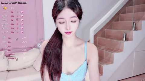 Media: A video of a young Asian woman with long brown hair in a ponytail, wearing a light blue lace bralette, standing in a modern, pink-themed living room with a white leather sofa and beige stairs.