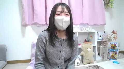 Media: Video of an Asian woman with straight black hair, wearing a white face mask, gray cardigan, and seated at a desk in a pastel-pink room with a toy bear and framed photos.