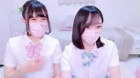 Media: Video of two young Asian girls in matching white school uniforms, wearing pink bows and face masks, sitting side by side in a bright, minimalist bedroom.