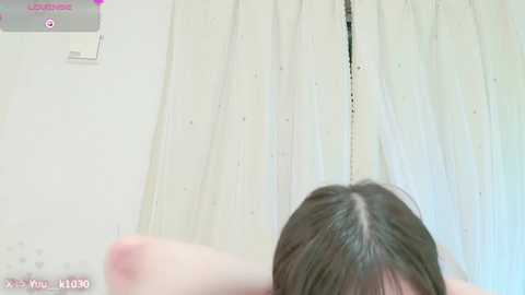 Media: A video of a woman with long, straight brown hair, seen from the back, standing in front of white, slightly textured curtains.