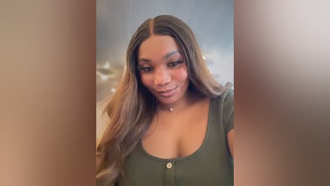 Media: Video of a Black woman with long, wavy brown hair, wearing an olive green top with a low neckline, smiling softly, indoors with blurred background.